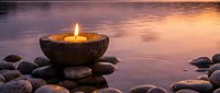 Stone bowl candle burning peacefully on stacked river rocks beside calm water at sunset creating serene spa meditation atmosphere for wellness relaxation.