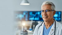 Senior male doctor with gray hair and glasses smiling confidently in modern medical facility with blue technology displays in background.
