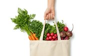 Hand holding a reusable cotton tote bag full of fresh organic vegetables including carrots, radishes, and beets, isolated on white.