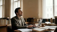 Young professional businessman working on laptop in bright office library with books, natural sunlight streaming through windows creating warm atmosphere.