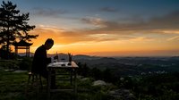 Man sitting on chair at wooden table on hillside overlooking valley at sunset, working on laptop, suitable for remote work, freelance, and business lifestyle concepts.