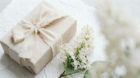 Elegant gift box wrapped in natural kraft paper with rustic twine bow alongside delicate white flowers on soft textured background for special occasions.