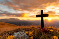 Wooden cross on mountain summit at dramatic sunset with golden light, orange clouds and mountain silhouettes in background, wild flowers in foreground.