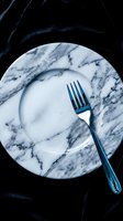 Empty marble plate with blue fork on dark textured background, overhead view. Minimalist table setting for fine dining, restaurant menu design, food photography, and culinary concepts.