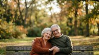 Senior couple embracing on wooden bench in autumn park, wearing glasses and casual clothing, enjoying outdoor time together in fall season.