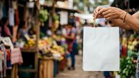 Person holding white blank shopping bag at outdoor market with blurred crowd and flower stalls in background for retail and commerce concepts.
