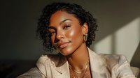 Young woman with curly hair and glowing skin in beige blazer illuminated by warm sunlight, showcasing natural makeup and gold jewelry.