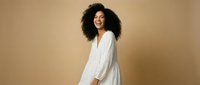 Young African American woman with curly hair wearing white flowing dress dancing joyfully against beige background for lifestyle and fashion content.