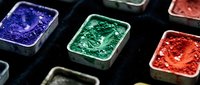 Colorful glitter and sequin containers arranged in rows on dark surface, featuring blue, green, and red shimmery materials for craft and design projects.