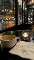 Coffee cup and candle on a cafe table with a napkin reading Sorry, rainy night city lights reflected on wet window glass.