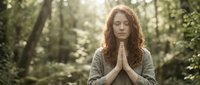 Peaceful young woman with curly red hair meditating in prayer pose outdoors in sunlit forest setting for mindfulness and spiritual wellness practices.
