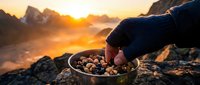 Hiker's hand reaching into bowl of trail mix at sunrise in mountains with misty peaks and golden sky in background.