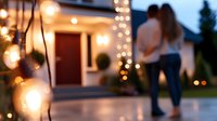 Romantic couple silhouette on patio with glowing string lights and bokeh effect creating intimate atmosphere at dusk, defocused background shows modern home exterior.