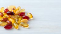 Colorful dietary supplement capsules and pills scattered on white wooden surface, featuring omega-3, fish oil, and vitamin E supplements in red, yellow and amber tones.