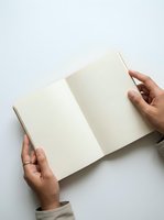 Person with medium skin tone holding open blank cream notebook with both hands against white background for writing, journaling, planning, or branding mockup purposes.