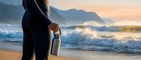 Surfer in wetsuit holding surfboard at sunrise beach with rolling ocean waves and misty mountains in background.