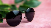 Classic aviator sunglasses with dark lenses on pink textured background with green leaves, selective focus creating soft dreamy mood for fashion and lifestyle content.