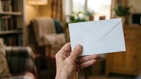 Senior man holding blank white envelope in home interior with warm lighting and bookshelves, suitable for mail, correspondence, and communication concepts.