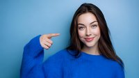 Young Caucasian woman with long dark hair wearing bright blue sweater pointing finger and smiling against blue background, expressing positive emotions and confidence.