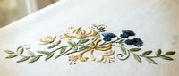 Delicate floral embroidery with blue flowers and green leaves on white linen fabric, showcasing traditional needlework craftsmanship for home decor projects.