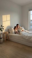 Mother and daughter reading book together on bed in bright minimalist bedroom with natural sunlight streaming through window creating warm atmosphere.