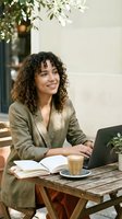 Young mixed-race businesswoman with curly hair working on laptop at outdoor cafe table with coffee and notebook, smiling while remote working in natural light.