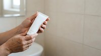 Close-up of mature man's hands holding blank white cosmetic tube in modern bathroom setting for skincare product mockup and advertising purposes.