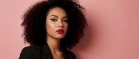 Young African American businesswoman with curly hair and red lipstick in black blazer against pink background for professional corporate branding.