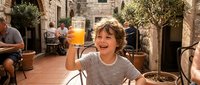 Young girl with brown hair enjoying fresh orange juice at outdoor restaurant terrace with stone architecture and Mediterranean atmosphere during family dining.