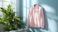 Pink formal shirt hanging on wall with natural window light and green plant, casting soft shadows. Minimalist fashion composition for business attire.