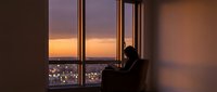 Silhouette of businessman working on laptop by large windows overlooking city lights at sunset, modern office interior with warm evening glow.