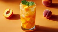 Fresh peach iced tea garnished with mint leaf in tall glass, surrounded by ripe cut and whole peaches on warm orange background, refreshing summer drink.