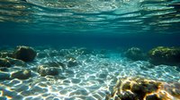 Underwater view of sunlight creating rippling patterns on sandy ocean floor through crystal clear turquoise water, with rocks and coral formations visible.