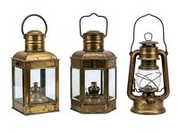 Vintage brass lanterns with glass panels and handles, featuring weathered patina finishes. Left and center lanterns have square frames with hinged doors, while right lantern displays classic hurricane design. All three isolated on white background, ready for design projects, home decor catalogs, or product mockups.