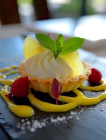 Mini lemon tart garnished with whipped cream, fresh pineapple, mint leaves and raspberries on black slate plate with decorative mango sauce swirls and powdered sugar.