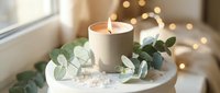 Lit white candle surrounded by eucalyptus leaves on marble surface with warm bokeh lights creating cozy spa atmosphere for relaxation and wellness.