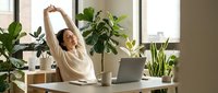 Young Asian woman stretching arms above head while working from home office with laptop and green plants, taking wellness break during remote work.