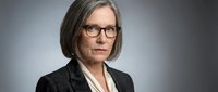 Middle-aged woman with shoulder-length gray hair and glasses wearing professional black blazer and white blouse against neutral gray background for corporate portraits.