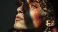 Young man with curly hair and facial piercings in dramatic colorful lighting with orange and blue tones creating artistic portrait for creative projects.