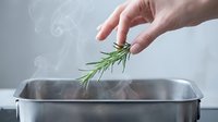 Hand adding fresh rosemary herb to stainless steel pot with steam, cooking ingredient preparation for culinary use.
