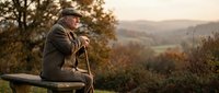 Elderly man with walking stick sitting on bench overlooking autumn countryside landscape at golden hour sunset, peaceful retirement moment.