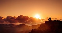 Person silhouette meditating on mountain peak during golden sunrise with dramatic clouds and layered mountain ranges creating peaceful spiritual moment.