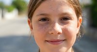 Young Caucasian girl with freckles smiling outdoors on sunny day. Natural portrait of happy child with brown hair in casual setting for family content.