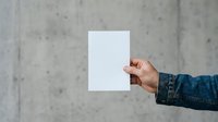 Person in denim jacket holding blank white paper against concrete wall background for mockup and branding purposes.
