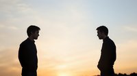 Dramatic silhouette of two men facing each other against golden sunset sky, depicting confrontation, rivalry or serious conversation between male figures.