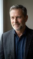 Mature man with gray hair and beard wearing dark blazer over navy shirt, studio portrait against neutral background for business and professional use.