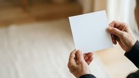 Mature adult male hands holding blank white card or letter against blurred indoor background, suitable for business communication, greeting cards, invitations, and marketing mockups.