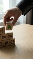 Businessman hand placing wooden cube with green house icon on eco-friendly blocks featuring renewable energy symbols on office desk for sustainability concept.