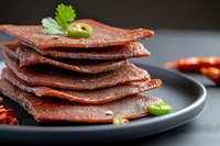 Spicy dried meat jerky snacks stacked on plate with chili peppers and cilantro garnish for protein-rich healthy eating and food photography.