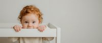 Adorable redhead toddler with curly hair peeking over white chair back, curious expression, neutral background for family and childcare concepts.